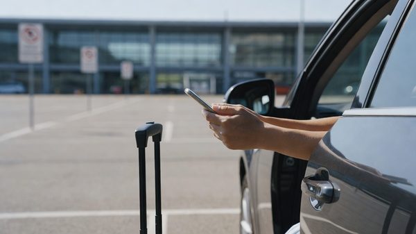 Traveler holding a phone beside a car in an airport parking lot with signage in the background